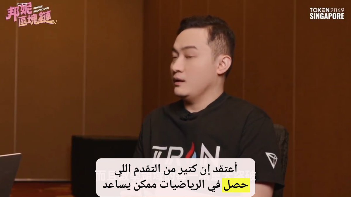 A man in a black 'TRAN' t-shirt being interviewed at TOKEN 2049 SINGAPORE, with Arabic subtitles translating his thoughts on sports achievements.