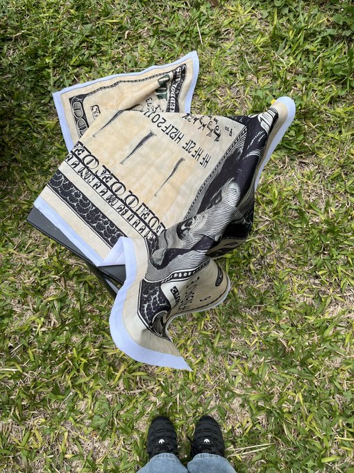 A crumpled US $100 bill rests on a grassy lawn, photographed from above with a person's black shoes and blue jeans visible at the bottom of the frame.