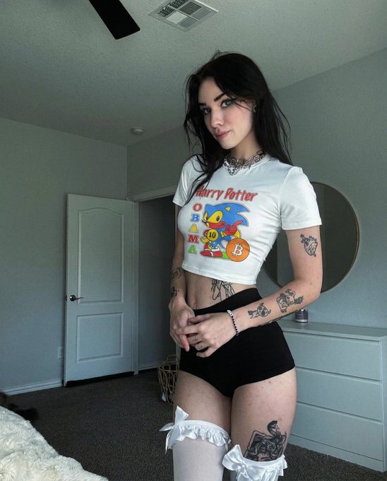 A young woman with dark hair and tattoos poses in a bedroom wearing a white "Harry Potter" graphic crop top, black shorts, and white thigh-high socks, saying "gm" (good morning) to her followers.