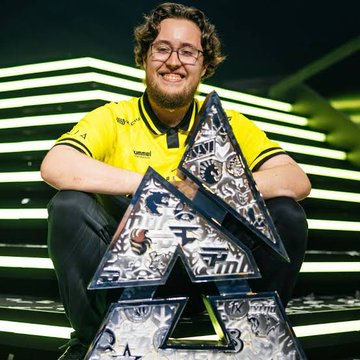 A smiling young man wearing glasses and a yellow and black jersey (likely an esports player) posing behind a large triangular trophy with intricate designs, on a stage with green lighting.