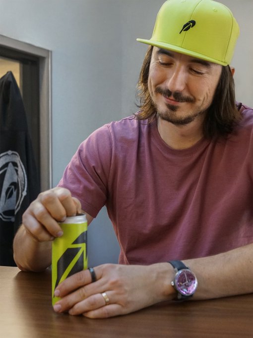 A man with long hair and a beard wearing a neon yellow cap and maroon t-shirt, opening a neon yellow and black beverage can at a table.