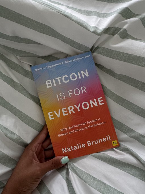 A hand holding the book 'BITCOIN IS FOR EVERYONE: Why Our Financial System is Broken and Bitcoin is the Solution' by Natalie Brunell against a striped bedsheet background.