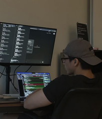 A person wearing a cap and glasses sitting at a desk with multiple monitors displaying what appears to be code or trading interfaces.
