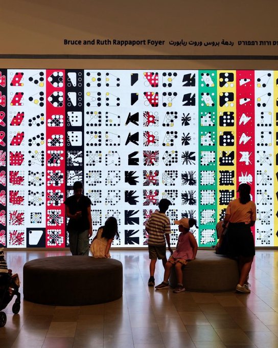 An indoor art installation in the 'Bruce and Ruth Rappaport Foyer' (text in English, Hebrew, and Arabic) featuring a large illuminated wall mural with colorful geometric and figurative patterns, observed by several visitors including children.
