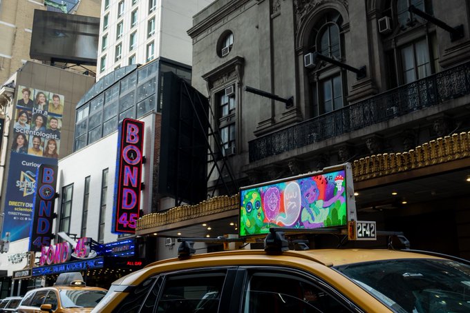 A yellow taxi cab passes in front of the Bond 45 restaurant and theater marquee in NYC, featuring a colorful digital billboard displaying artwork that aligns with @Artcrush's mission of showcasing defining artists in iconic city locations.