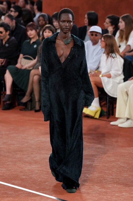 A fashion runway photo of a model with a short buzz cut wearing a long, dark green unbuttoned velvet coat dress with a necklace, walking down a terracotta-colored catwalk with seated audience members visible behind.