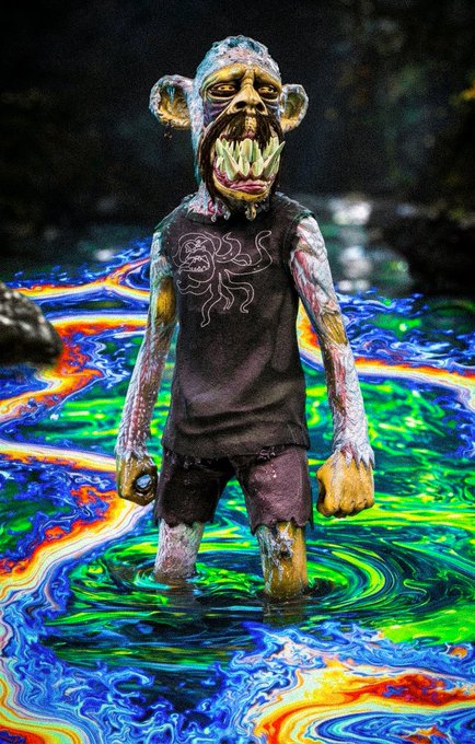 A grotesque mutant ape figurine with an oversized fanged mouth stands in a surreal pool of swirling rainbow liquid, evoking the bizarre aesthetic of the Mutant Ape Yacht Club NFT collection in celebration of its 5-year anniversary. The ape wears a black sleeveless shirt featuring a small illustrated creature, tying into the diverse trait variations that made the collection iconic.
