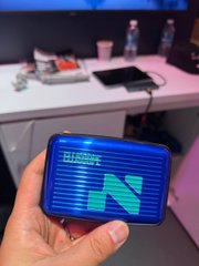 A hand holds a smartphone displaying a dark screen with a glowing blue 'N' logo, set in what appears to be a tech or gaming event environment with monitors in the background.