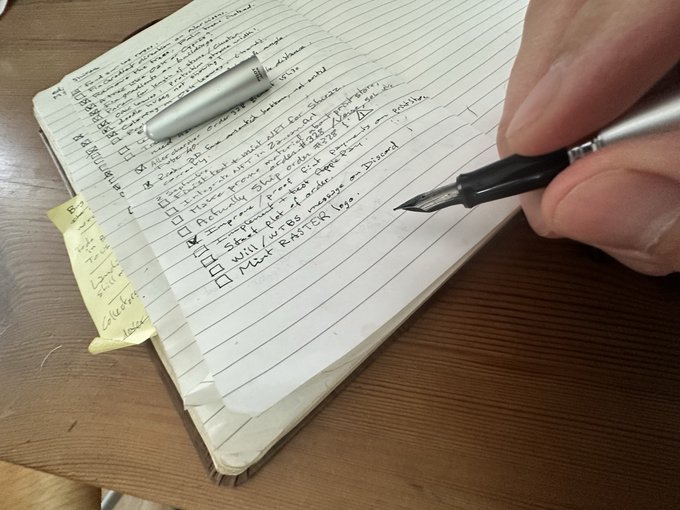 A hand holding a fountain pen writes handwritten notes and a checklist on a lined notebook page with a yellow sticky note attached.