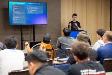 A presenter stands at a podium in front of a large screen displaying code, addressing an audience at what appears to be a technical conference.