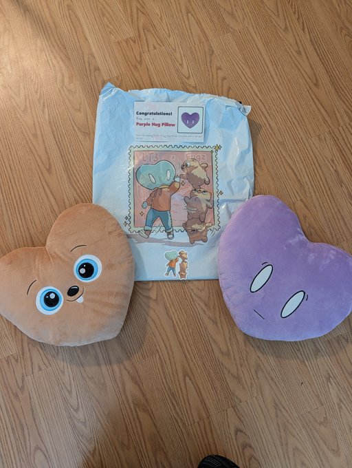 Two heart-shaped plush pillows—one orange with a Stitch face and one purple—rest on a wooden floor next to their original packaging labeled 'Purple Hug Pillow'.