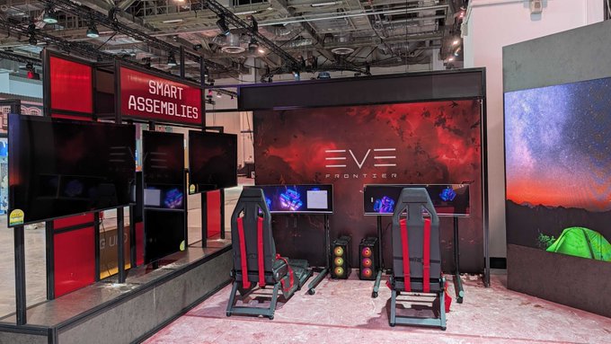 A trade show booth displaying 'SMART ASSEMBLIES' and 'EVE FRONTIER' branding, featuring gaming/VR chairs with red accents, computer monitors, and a large screen showing a space/galaxy scene.