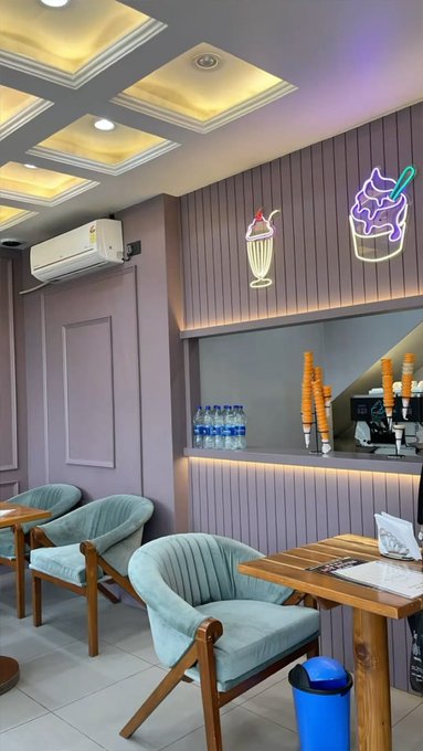 The interior of a dessert shop features mint green chairs, wooden tables, ice cream cone decorations, and neon signs on a wood-paneled wall.