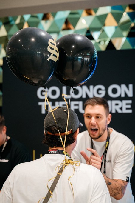 Two men celebrate in front of a "GO LONG ON THE FUTURE" backdrop, with one holding two black balloons displaying the Bitcoin '₿' symbol.