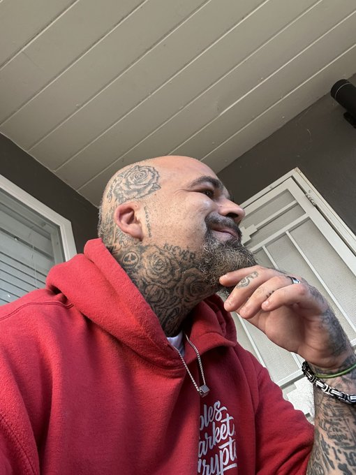 A selfie of a heavily tattooed bald man with a beard, wearing a red hoodie, standing outside a house.