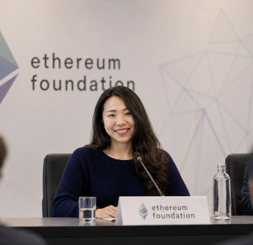 The image shows a woman sitting at a table, smiling for the camera while wearing a blue sweater. The table has a nameplate that reads "Ethereum Foundation", indicating that she might be attending an event related to the foundation or its projects. There are two other people in the background who appear to be engaged in conversation as well. A water bottle is also visible on the table, suggesting that refreshments may have been provided during the event.
