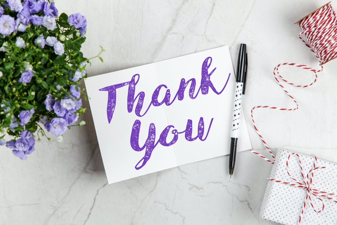 A 'Thank You' greeting card with purple glittery lettering displayed on marble alongside purple flowers, a pen, and a wrapped gift with red and white twine.