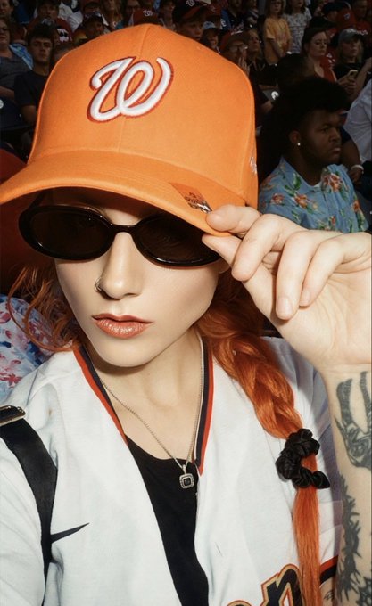 A person with orange hair wears an orange Washington Nationals 'W' baseball cap and dark sunglasses, posing with their hand near their face at what looks like a sporting event.