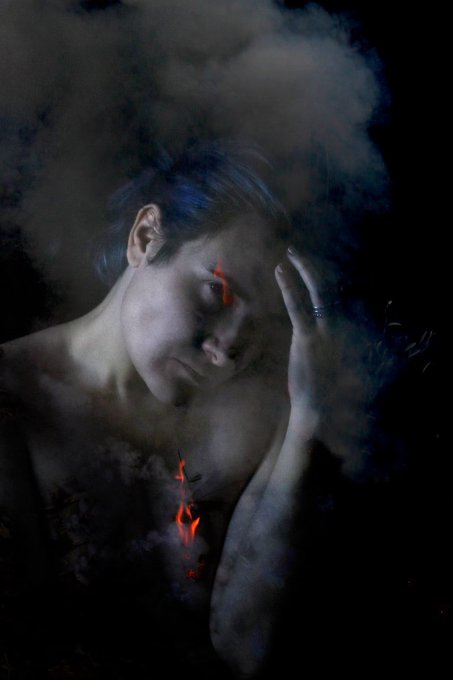 Artistic portrait of a shirtless figure with blue-tinted hair, eyes closed, set against a dark background with swirling clouds and glowing red lava-like accents flowing from the eyes and chest.