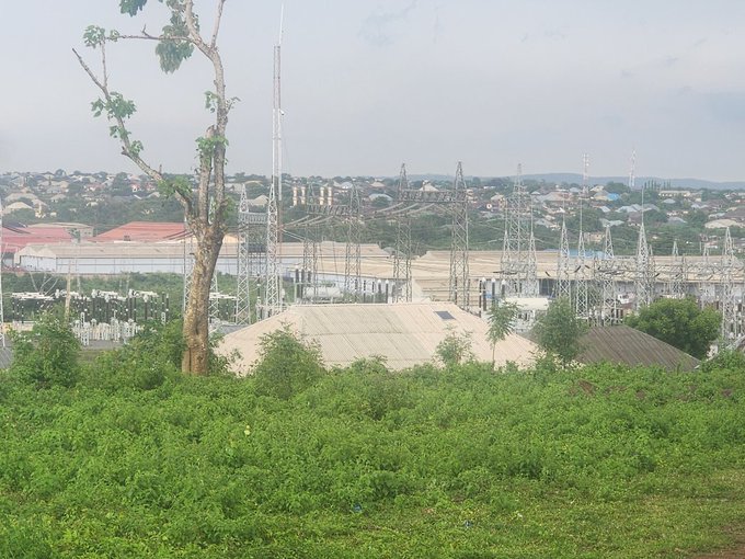 A landscape view showing an electrical substation with transmission towers and power lines, surrounded by green vegetation and buildings in the background.