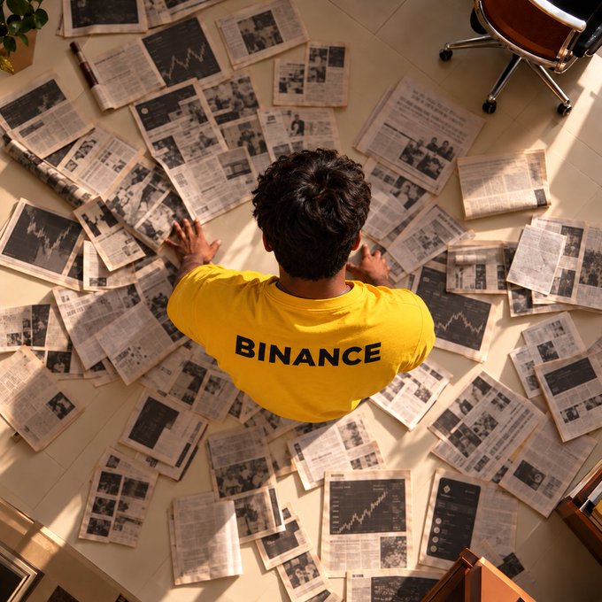 Overhead view of a person in a yellow "BINANCE" shirt crouching amid a chaotic floor covered in scattered financial newspapers and profit charts, illustrating the overwhelming feeling of drowning in market news and trading screenshots.