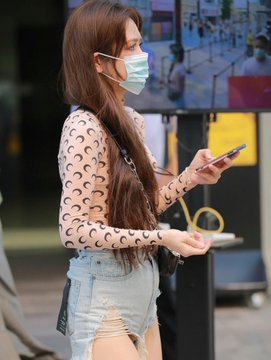 A young woman wearing a sheer black patterned long-sleeve top, light blue denim shorts, and a surgical mask stands on a city street while looking down at her phone. The post suggests the "unexpected gain" spotted during a casual night stroll is this stylish passerby.