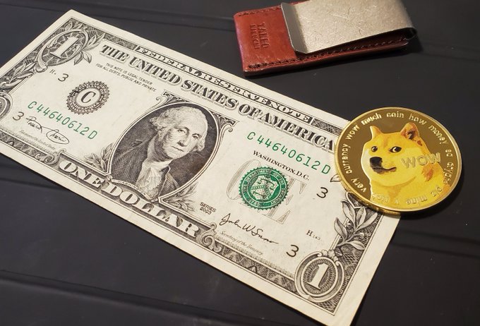 A U.S. one-dollar bill placed next to a gold-colored Dogecoin commemorative coin on a dark surface, with a brown leather wallet partially visible in the upper portion of the frame.