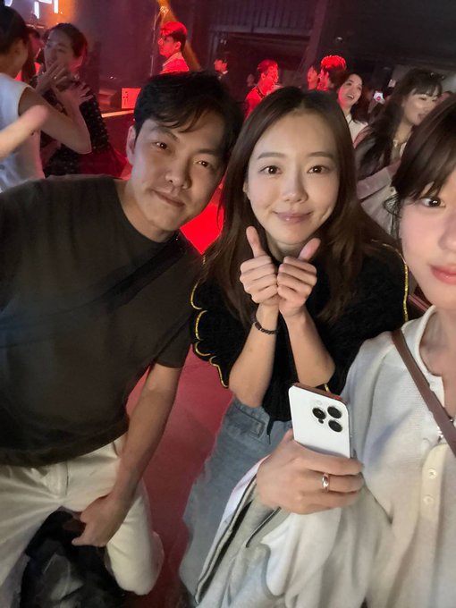 Three fans smile for a selfie at what appears to be a VIP event, with the central woman making a finger heart gesture while holding an iPhone, capturing the excitement of meeting the personality "Jadoodoo" mentioned in the caption. The dimly lit venue with red-toned lighting in the background suggests an evening event, consistent with the social gathering referenced in the post.