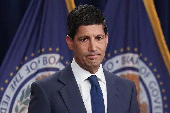 A man in a dark suit and blue tie stands before two "Board of Governors of the Federal Reserve System" flags, appearing solemn as he addresses the press. The image accompanies breaking news of the Federal Reserve raising interest rates by 25 basis points to a range of 3.75%–4.00%.
