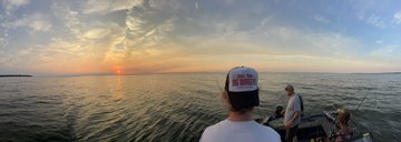 A scenic photograph taken from a boat at sunset, showing two people looking out over calm water towards the horizon where the sun is setting, with dramatic clouds overhead.