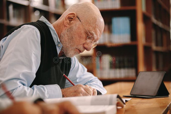 An elderly bald man with glasses, wearing a light blue shirt and black vest, writing with a red pencil at a desk with a tablet and books, set against a library background.