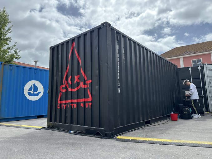 The image features a large black shipping container with red graffiti on its side, which has been turned into an art installation. The container is situated next to a building, possibly a house or a warehouse. A person can be seen standing near the container, likely observing and appreciating the artwork. In addition to this, there are two boats visible in the image - one located at the top left corner of the frame and another further down on the right side.

The post accompanying the image is titled "@basseadx @CultCryptoArt @NFCsummit sailing the @opensea". This post seems to be promoting an art project or event related to shipping containers, graffiti, or sailing. The combination of the large black container with red graffiti and the boats in the background creates a visually interesting scene that connects the artwork to the surrounding environment.