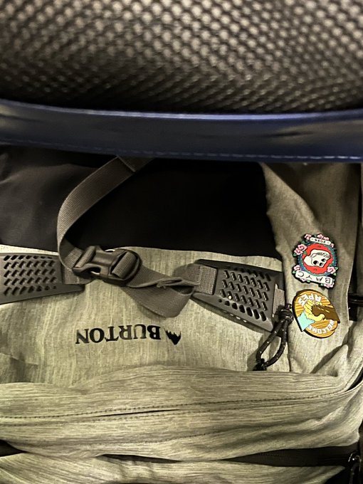 A close-up of a gray Burton backpack with visible buckles, strap, mesh panel, and decorative pins attached to the side.