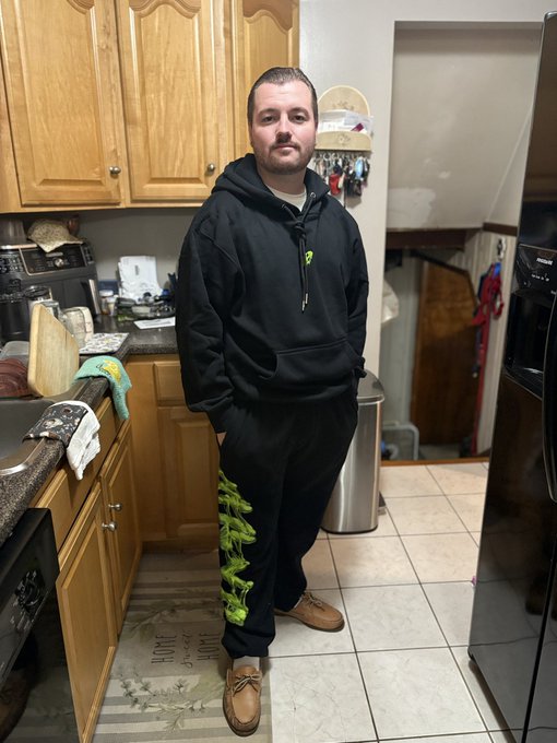 A man wearing a black hoodie and matching sweatpants with green graphics stands in a kitchen with wooden cabinets and a black refrigerator.