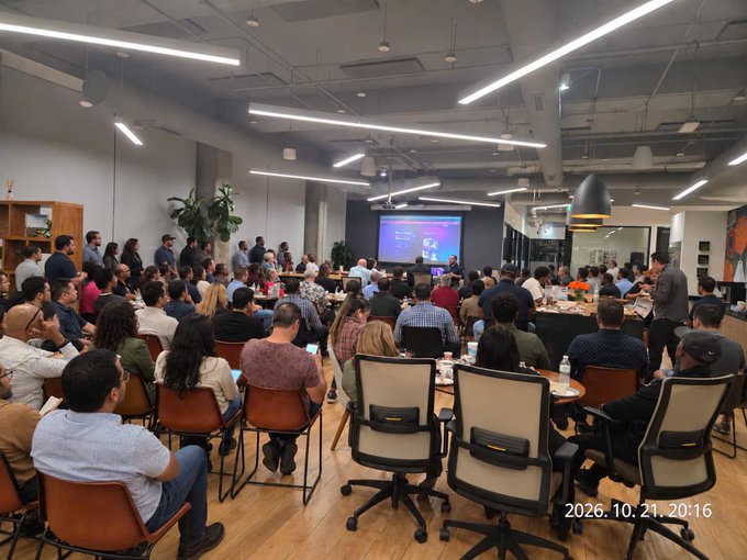 Photograph of a crowded office presentation with attendees seated around tables facing a large screen; a timestamp overlay reads “2026.10.21 20:16.”