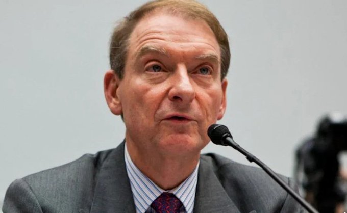 A close-up of an older man in a gray suit and red patterned tie speaking into a microphone.