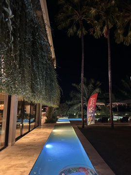 A nighttime view of a resort pool area with palm trees, illuminated blue water, lounge chairs, and a red flag reading 'SANDALS'.
