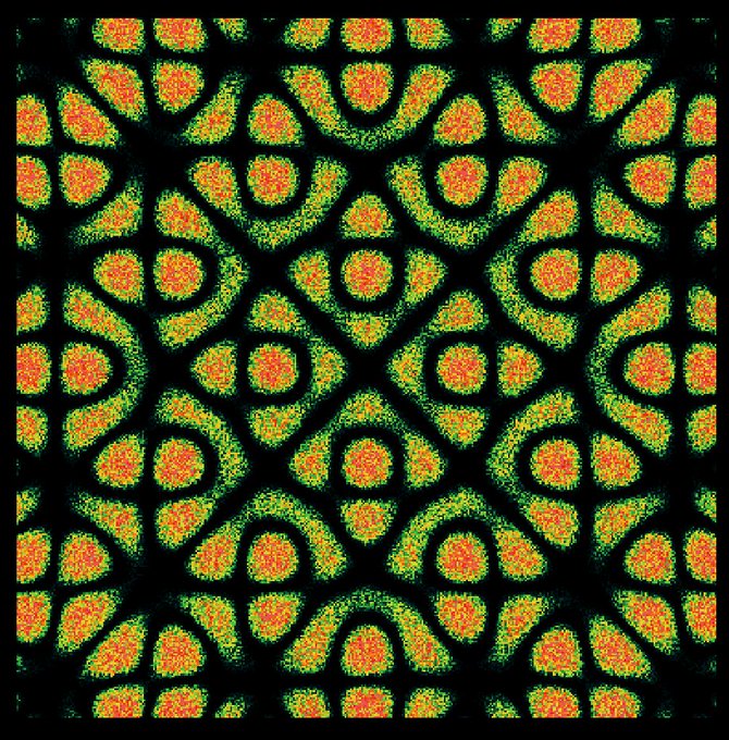A symmetrical abstract pattern featuring orange and green shapes arranged in a kaleidoscopic design on a black background, resembling organic or cellular forms.
