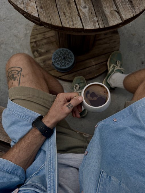 an overhead shot of two people sitting at a wooden spool table, one holding a coffee mug and the other wearing green sneakers, with a small foot tattoo visible.