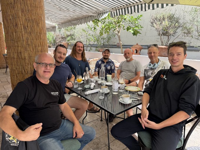 A group of seven men sit around an outdoor dining table on a patio under a striped awning, enjoying a meal together.