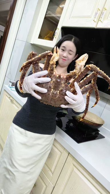 A woman wearing white gloves holds up a massive king crab in a kitchen.
