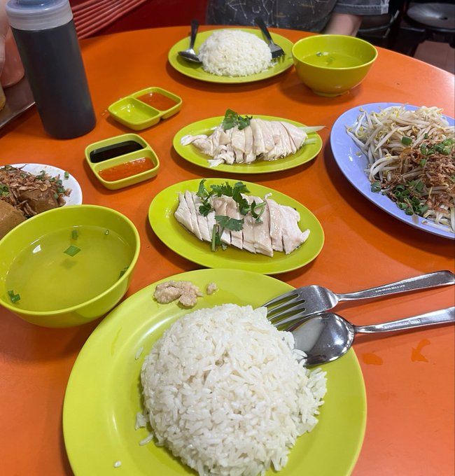 Two servings of Hainanese chicken rice with steamed white rice, sliced chicken garnished with cilantro, a bowl of clear broth, bean sprouts, and chili sauce are spread across an orange table, exemplifying the classic Singaporean hawker fare the post recommends seeking out during Token2049—even if the staff are famously rude.