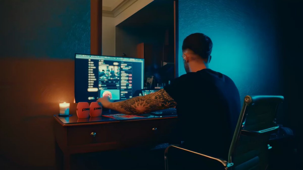 A person with a tattooed arm sits in a dimly lit room with blue lighting, working at a computer displaying what appears to be a game interface, with a candle and two small figurines on the desk.