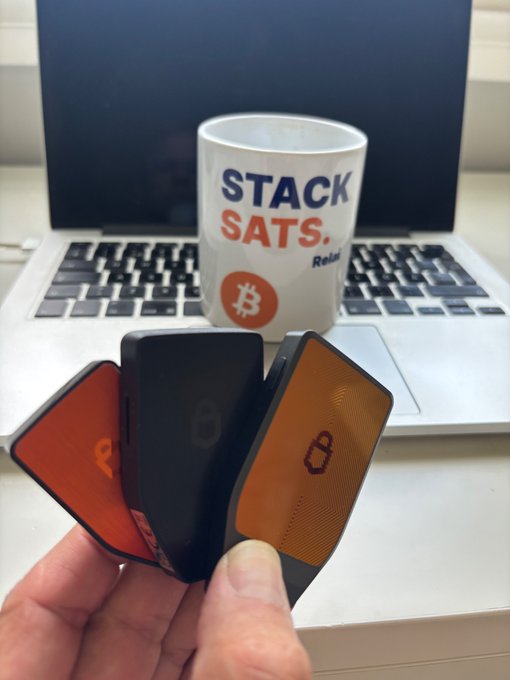 A hand holds three crypto hardware wallets (in black and orange) in front of a laptop and a white mug featuring Bitcoin's logo and the text 'STACK SATS. Relax'.