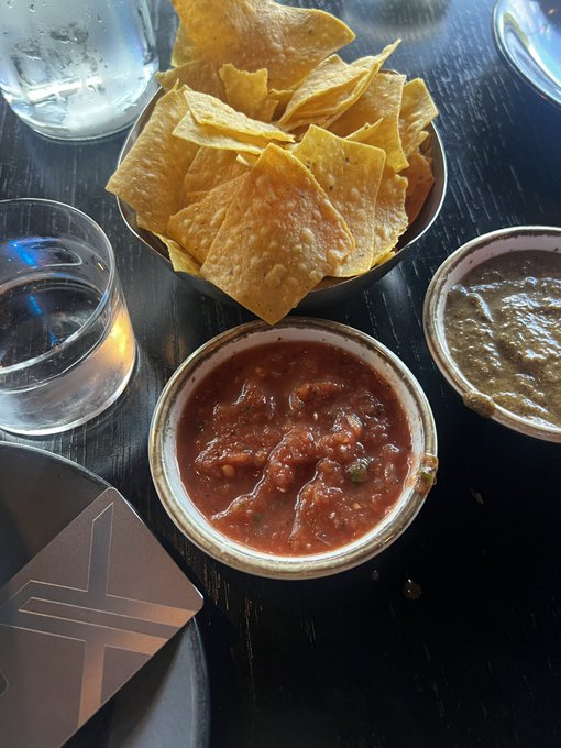 A bowl of tortilla chips accompanied by salsa and guacamole sits on a dark wooden table alongside water glasses and a coaster featuring the X logo, seemingly enjoyed as a celebratory lunch following a transaction on X Money.