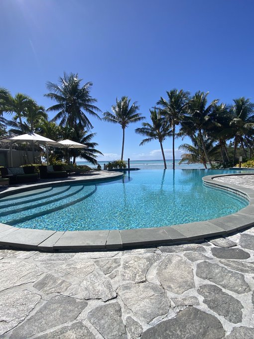 A beautiful resort pool with crystal-clear blue water, surrounded by palm trees and stone paving, overlooking a calm sea under a clear blue sky.