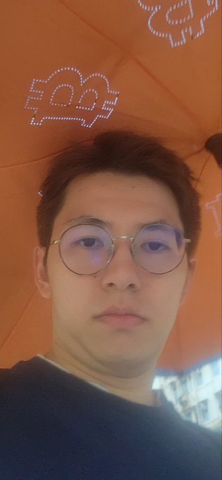 A selfie-style photo of a young man wearing round wire-frame glasses under an orange umbrella decorated with cryptocurrency-themed pixel art symbols including an Ethereum-style logo.