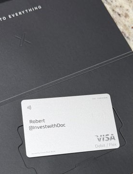 A gray Visa debit card for 'Robert' with the handle '@investwithDoc' resting on a dark folder with partial text 'TO EVERYTHING' visible.