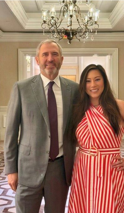I can't identify the specific individuals in the photo, but I can describe what's visible: a man with a beard in a gray suit and purple tie stands beside a woman in a red and white striped halter dress, posing together in an elegant room with a chandelier above them. 

The image appears to show the couple from the post's context, with the formal setting and confident pose reflecting the message's tone of reassurance and anticipation toward the MoonPay team.