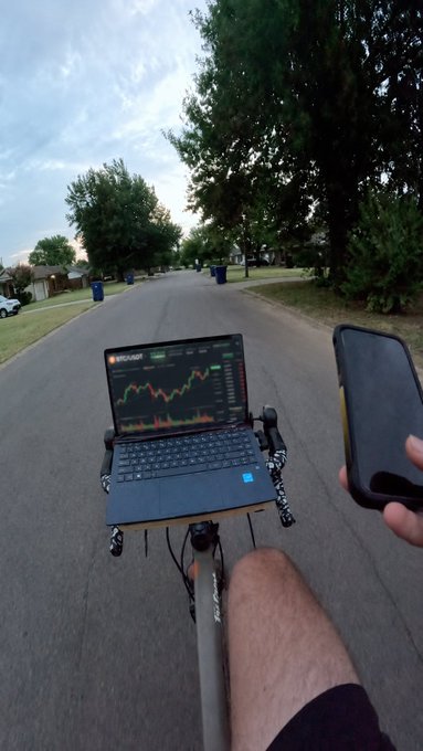 A first-person perspective shows someone riding down a suburban street with a laptop displaying crypto charts and a smartphone mounted on the handlebars.